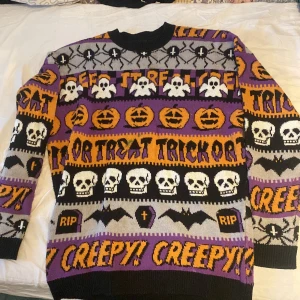 Halloweentröja med dödskallar och pumpor från FSBN - Säljer en supercool Halloweentröja från FSBN i storlek M. Den är svart med ett färgglatt mönster av dödskallar, pumpor, fladdermöss och text som 'Trick or Treat' och 'Creepy!'. Perfekt för höst och Halloweenfester! Tröjan är stickad och har långa ärmar. Grymt tillfälle att sticka ut på festen! 🎃💀den har en grön fläck ):