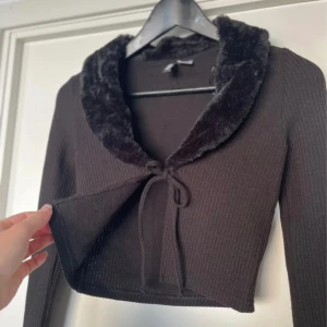 Svart ribbad kofta med fuskpäls - Säljer en superfin svart ribbad kofta med en mysig fuskpälsdetalj runt kragen. Den är croppad och har långa ärmar, perfekt för höst och vinter. Koftan knyts framtill med ett sött band. Passar både till vardags och fest! 🖤Passar XXS-XS