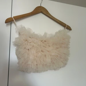 Beige volangtop från Gina Tricot - Säljer en superfin beige volangtop från Gina Tricot. Den är axelbandslös och har en fluffig volangdesign som ger en riktigt söt look. Perfekt för fest eller sommarhäng! Toppen är i nyskick och väldigt stretchig passar definitivt S-L