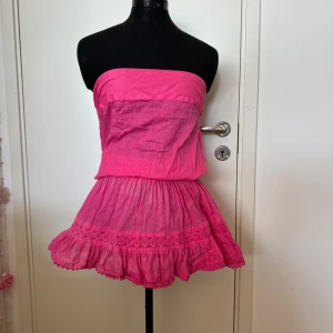 Rosa axelbandslös klänning - Säljer en superfin rosa axelbandslös klänning med volangdetaljer. Den är perfekt för sommaren och har en elastisk midja för en bekväm passform. Klänningen är kort och har en söt spetsdetalj längs fållen. Perfekt för en dag på stranden eller en sommarfest! 🌸