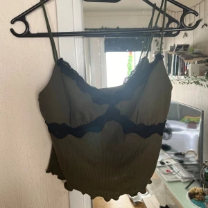 Olivgrön ribbad topp med spetsdetaljer - Säljer en superfin olivgrön ribbad topp med svarta spetsdetaljer. Toppen är ärmlös och har en croppad passform, perfekt för sommaren eller en festkväll. Den har tunna axelband och en söt spetskant vid halsringningen. Passar perfekt till både jeans och kjol!
