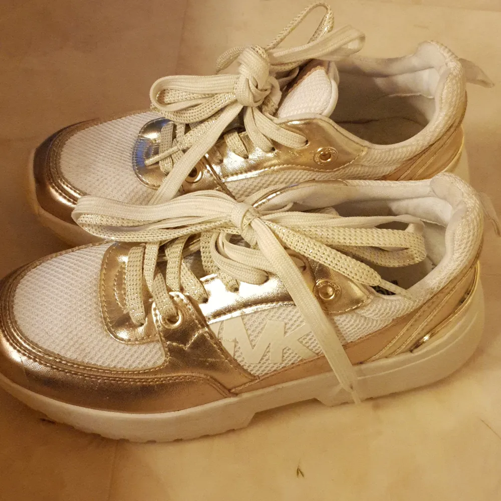 Säljer ett par snygga sneakers från Michael Kors i vitt och guld. Skorna har en glittrig finish med snörning och en bekväm sula. Perfekta för att ge din outfit lite extra bling! Stora i storleken. . Skor.