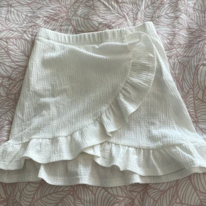 Vit omlottkjol med volanger - Säljer en superfin vit omlottkjol från märket 157. Kjolen har en söt volangdetalj längs kanten och är perfekt för sommaren. Den är i storlek 150 och passar bra för både vardag och fest. Materialet känns lätt och luftigt, vilket gör den bekväm att bära hela dagen. Perfekt att matcha med en enkel topp eller blus!