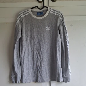 Grå långärmad tröja från Adidas - Säljer en snygg och bekväm grå långärmad tröja från Adidas. Tröjan har de klassiska tre ränderna längs ärmarna och Adidas-loggan broderad på bröstet. Tröjan är i bra skick och gjord av mjukt bomullsmaterial. 