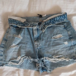 Blå jeansshorts från Divided - Säljer ett par blå jeansshorts från Divided. De är högmidjmidjade men kan även vikas ner till låg midja och har en sliten look med flera revor och slitningar. Perfekta för sommaren och ger en cool och avslappnad stil. Shortsen har två fickor fram och två bak, samt en dragkedja och knapp framtill. (pris kan diskuteras)