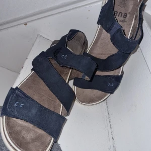 Blå sandaler från Jana - Snygga och bekväma blå sandaler från Jana med breda remmar och kardborreknäppning. Perfekta för sommarens varma dagar. Sandalerna har en ljus sula som ger en fin kontrast till det blå materialet.