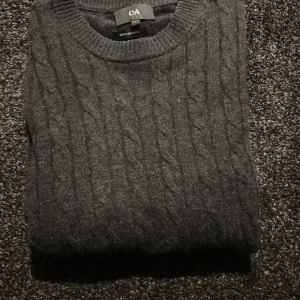 Stickad Kashmir  tröja  - Säljer en stilren grå kabelstickad tröja från C&A, tillverkad med kashmir för extra mjukhet. Tröjan har en klassisk rund hals och långa ärmar, perfekt för kyligare dagar. Passar både till jeans och mer uppklädda tillfällen.