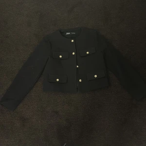 Svart tröja från Zara - Snygg svart kavaj från Zara med guldknappar och två fickor framtill. Perfekt för en stilren look. Kavajen har en klassisk design med långa ärmar och rund halsringning.