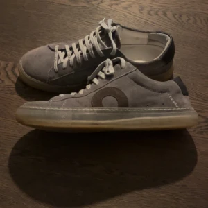 Grå sneakers från Oliver Cabell - Snygga grå sneakers i mocka från Oliver Cabell med en genomskinlig sula. Skorna har en stilren design med en diskret logga på sidan och är perfekta för en avslappnad look. Hör av dig vid frågor eller funderingar. Pris ej hugget i sten 