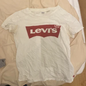 Vit t-shirt från Levi's - Säljer en klassisk vit t-shirt från Levi's med det ikoniska röda logotyptrycket på framsidan. Perfekt för en avslappnad stil. T-shirten är kortärmad och tillverkad i bomull.