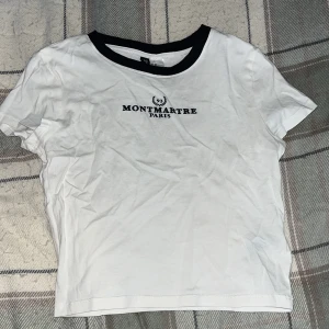 Vit magtröja med tryck från H&M Divided - Säljer en vit magtröja från H&M Divided med svart text 'Montmartre Paris' på bröstet. T-shirten har en rund halsringning med svart kant och korta ärmar. Perfekt för en avslappnad stil.