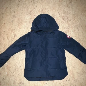 Canada Goose jacks - Säljer en mörkblå dunjacka från Canada Goose med huva och det klassiska märket på ärmen. Jackan är perfekt för kalla dagar och har en stilren design. Jackar funkar även i lite varmare väder då den passar till många outfits. Säljer jackan billigt eftersom de lilla hålet på ryggen. Man märker det knappt. Orginal påsen tillkommer ochså vid köp 