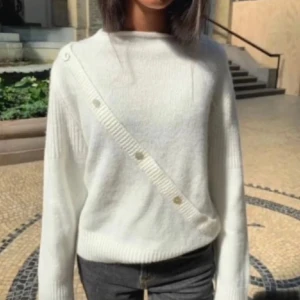 Vit stickad tröja med knappar - Design by Si. Säljer en stilren vit stickad tröja med unika knappdetaljer i omlott-design. Tröjan har långa ärmar och en bekväm passform, perfekt för kyligare dagar. Ett måste i garderoben för en klassisk look. Helt slutsåld!