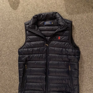 Ralph Lauren väst - Snygg mörkblå dunväst från Polo Ralph Lauren med dragkedja och broderad logga i rött på bröstet. Perfekt för våren som kommer nu och ger en stilren look. Västen har en hög krage och två fickor med dragkedja.