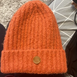 Orange stickad mössa från Pepe Jeans - Snygg orange stickad mössa från Pepe Jeans med en ribbad design. Mössan har en liten metallbricka med varumärket på framsidan. Perfekt för att hålla värmen under kyliga dagar.