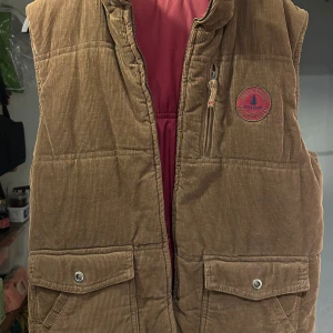 Vändbar manchesterväst - Säljer en vändbar väst med en sida i brun manchester och den andra i vinröd polyester. Västen har en dragkedja och två fickor med knappar på manchestersidan samt två dragkedjefickor på den vinröda sidan. Perfekt för att variera stilen!