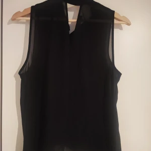 Svart ärmlös blus - Snygg svart ärmlös blus i ett lätt och luftigt material. Blusen har knappar framtill och en klassisk krage, perfekt för en stilren look.