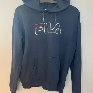 Mörkblå hoodie från FILA - Säljer en snygg mörkblå hoodie från FILA med deras ikoniska logga i vitt och rött på bröstet. Tröjan har en klassisk känguruficka och justerbar huva med dragsko. Den är i ett väldigt bra skick och har sätten används.