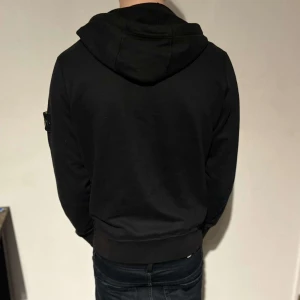 Stone island - Säljer en stilren svart huvtröja från Stone Island med dragkedja och deras ikoniska märke på ärmen.  Skick 9/10 använd runt 5 gånger. 