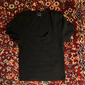 Svart kortärmad topp från Gina Tricot - Snygg svart topp från Gina Tricot med korta ärmar och v-ringning. Toppen har knappar framtill som en fin detalj. Perfekt för en stilren look.