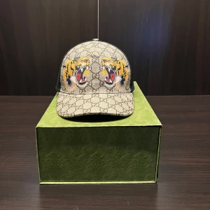 Gucci Keps - Säljer en snygg Gucci keps med ett tigerbroderi framtill. Kepsen har ett klassiskt mönster i beige och svart med en justerbar rem baktill. Perfekt för att ge din outfit en unik touch.