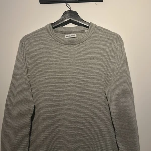 Grå stickad tröja från Jack & Jones - Säljer en stilren grå stickad tröja från Jack & Jones. Tröjan har en rund halsringning och långa ärmar, perfekt för kyligare dagar. Den är enkel att matcha med olika outfits och ger en avslappnad men ändå snygg look.