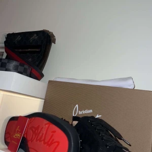 Svarta sneakers med nitar från Christian Louboutin - Snygga svarta sneakers från Christian Louboutin med ikoniska röda sulor och nitar på ovansidan. Skorna har snörning och är tillverkade i mocka. Perfekta för att ge en edgy touch till din stil.