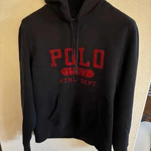 Svart hoodie från Polo Ralph Lauren - Säljer en svart hoodie från Polo Ralph Lauren med röd text 'POLO 1967 ATHL. DEPT.' på framsidan. Tröjan har en klassisk huva med dragsko och en stor magficka. Perfekt för en avslappnad stil.