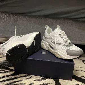 Vita och grå sneakers från Dior - Snygga och moderna sneakers från Dior i vitt och grått. Skorna har en tjock sula och är tillverkade i en kombination av syntetmaterial och mesh för en sportig look. De har snörning framtill och en diskret Dior-logga på sidan. Priset kan diskuteras 