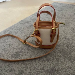 Beige och brun axelväska med röda detaljer - Snygg och kompakt axelväska i beige och brunt med röda detaljer. Väskan har en justerbar axelrem och guldfärgade spännen. Perfekt för att bära det nödvändigaste med stil.