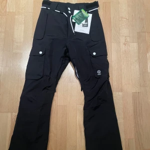 Skidbyxor Dope - Snygga svarta Skidbyxor från Dope med praktiska fickor och vita knappar. Perfekta för vinterns äventyr med en stilren design och bekväm passform. Byxorna har en hög midja och justerbar snörning i midjan för bästa komfort. Aldrig använda med lappar kvar.