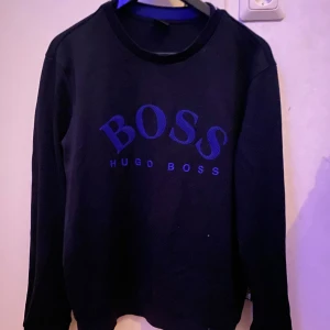 Svart tröja från Hugo Boss - Snygg svart tröja från Hugo Boss med stort BOSS-tryck på framsidan. Tröjan har en klassisk rund hals och långa ärmar, perfekt för en stilren look. Tröjan är svår och hitta nu förtiden, inte trasig nånstans🩵