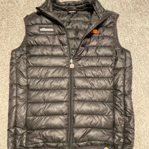 Svart Ellesse Väst - Snygg svart dunväst från Ellesse med dragkedja framtill. Västen är stilig och är perfekt för den kommande våren! Den har en broderad logotyp på bröstet och en hög krage för extra värme. Skick: 9,5/10, använt väldigt varsamt och försiktigt.