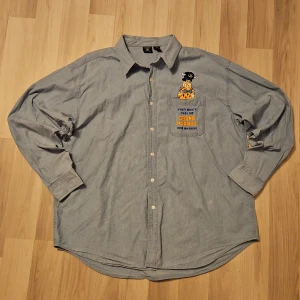 Blå skjorta med broderad figur från Warner Bros. - Säljer en blå skjorta från Warner Bros. med en broderad flintstones figur på bröstfickan. Storleken säger XXL men den passar som XL