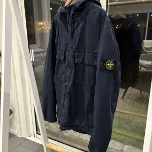 Mörkblå jacka med huva från Stone Island - Säljer en snygg mörkblå jacka från Stone Island med huva och dragkedja. Jackan har långa ärmar och en ikonisk logga på ärmen. Perfekt för en stilren look.
