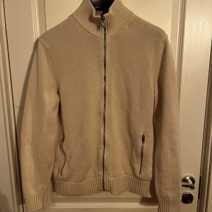 Beige stickad tröja från mango - Säljer en stilren beige stickad tröja från MNG med dragkedja framtill. Tröjan har en hög krage och långa ärmar, perfekt för kyligare dagar. Den har praktiska fickor med dragkedja på sidorna. Passar bra till både jeans och chinos. Fläck andra bild därav pris. Ingen fläck man tänker på. 