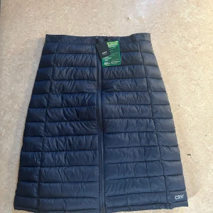 Marinblå täckkjol från CRW - Säljer en marinblå täckkjol från CRW, perfekt för kyliga dagar. Kjolen har en quiltad design med dragkedja framtill och elastisk midja för extra komfort. Den är både stilren och funktionell, idealisk för höst- och vinterbruk.