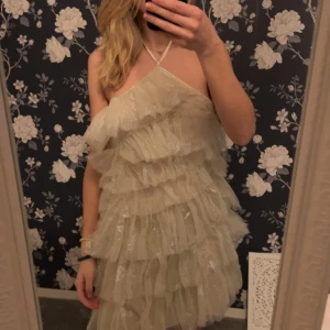 Glittrig halterneck klänning - Säljer en glittrig klänning med volanger och halterneck-design. Klänningen är i en ljus färg och har en kort längd. Aldrig använd - lappen finns kvar