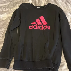 Svart tröja från Adidas med rosa logga - Säljer en svart tröja från Adidas med en stor rosa logga på framsidan. Tröjan har långa ärmar och ribbade muddar vid ärmslut och nederkant. Perfekt för en sportig look eller avslappnad stil. 🖤💗