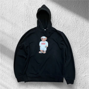 Svart hoodie med en supreme Björn på - Säljer en svart hoodie med ett coolt nallemotiv med supremes märke på framsidan. Hoodien har en klassisk passform med ficka på magen och en justerbar huva. Jag rekommenderar den starkt! Mitt pris är 199kr men om du blir eller är en följare så får du den för bara 179kr! Ställ gärna frågor!