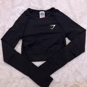 Svart croppad tröja från Gymshark - Säljer en snygg och sportig svart croppad tröja från Gymshark. Perfekt för träning eller en casual look. Tröjan har långa ärmar och ett diskret mönster som ger extra stil. Den är i mycket bra skick och redo för nya äventyr!