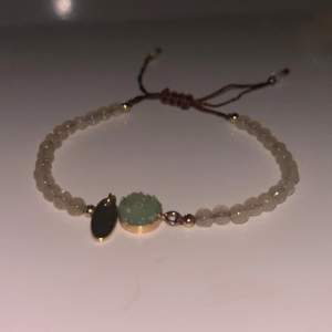 Snyggt armband med små pärlor och en grön detalj i mitten. Armbandet har justerbar knytning och en liten guldpläterad berlock. Perfekt för att ge en subtil touch av färg till din outfit.