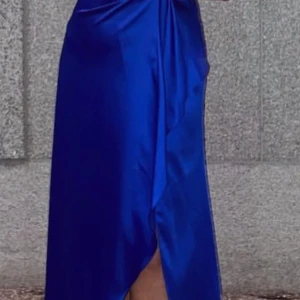 Blå axelbandslös klänning med bälte - Säljer en elegant blå axelbandslös klänning i satin med ett matchande bälte. Perfekt för festliga tillfällen med sin långa längd och draperade design. Klänningen har en vacker glans och en smickrande passform. använd endast en gång i några få timmar och har inga defekter