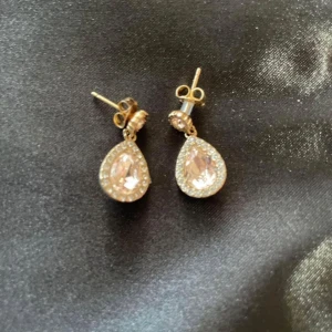 Guldörhängen med droppformade stenar - Säljer ett par eleganta guldörhängen med stora droppformade stenar omgivna av mindre glittrande stenar. Perfekta för att ge en lyxig touch till din outfit. Passar både till fest och vardag. Fästs med klassisk stift och plopp.