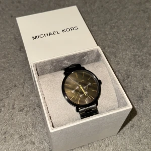 Svart klocka från Michael Kors - Säljer en stilren svart klocka från Michael Kors i mycket nyskick. Klockan har en svart urtavla med guldiga detaljer och en matchande svart länkarmband. Perfekt för både vardag och fest! Kommer med originalförpackning och etikett.