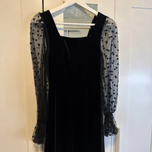 Svart sammetklänning med prickiga ärmar - Säljer en elegant svart sammetklänning med långa, transparenta ärmar i prickigt mönster. Perfekt för festliga tillfällen! Klänningen har en klassisk och stilren design som passar både höst och vinter.