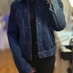 Mörkblå jeansjacka - Säljer en snygg mörkblå jeansjacka i bra skick. Den har en klassisk design med knappar framtill och två bröstfickor. Perfekt för vår och höst, och funkar till nästan alla outfits. Jackan har långa ärmar och en normal passform. Perfekt för dig som vill ha en stilren look!