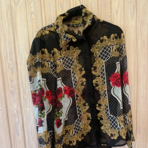 Mönstrad blus från Bik Bok - Säljer en superfin blus från Bik Bok i storlek XS. Den har ett unikt mönster med röda rosor och guldiga detaljer på en svart bakgrund. Perfekt för en festlig kväll eller en stilfull dag på stan. Blusen har långa ärmar och knappar framtill. Passar perfekt till både jeans och kjol!