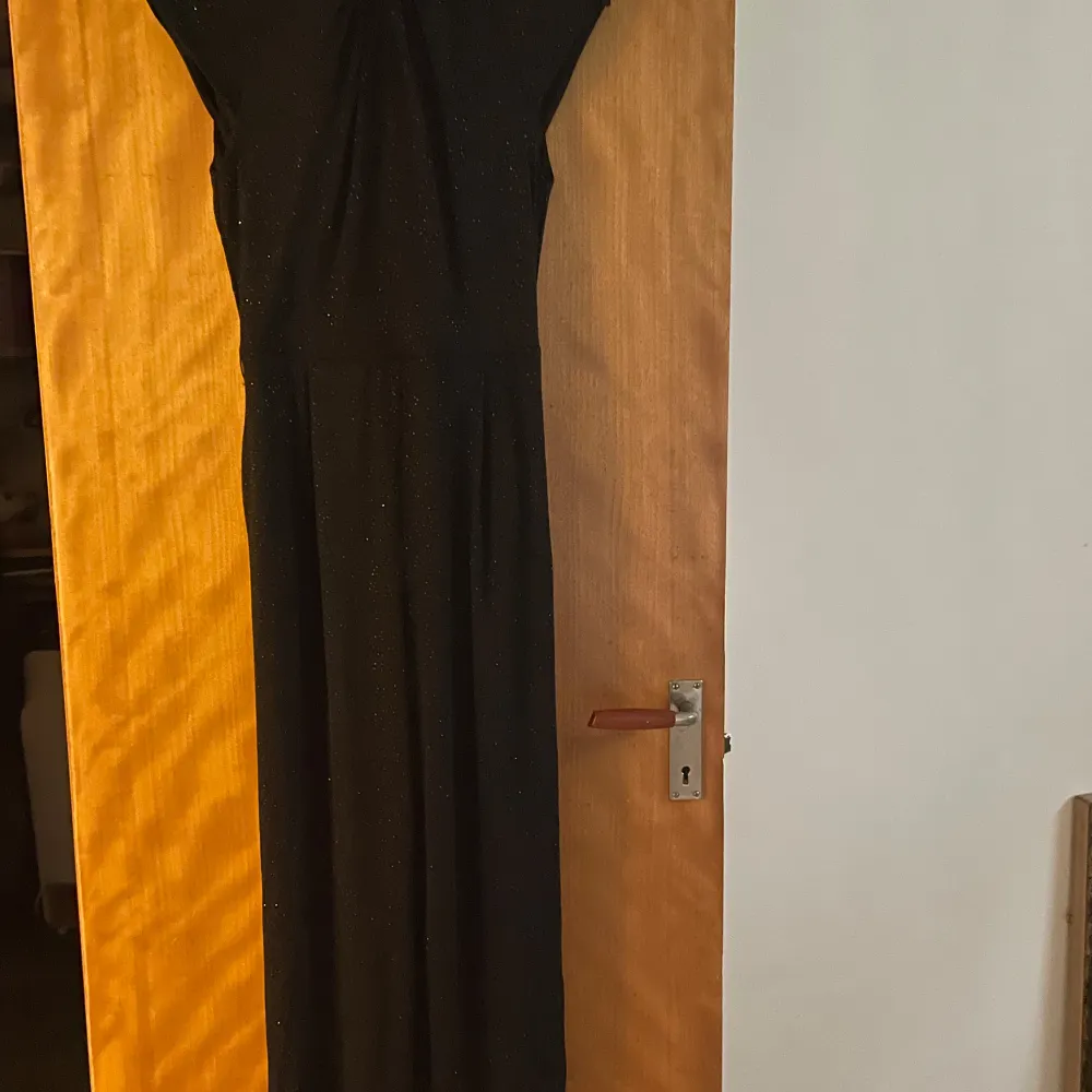 Snygg svart jumpsuit med glittrigt material, perfekt för festliga tillfällen. Den har en elegant passform och är ärmlös, vilket gör den idealisk för både nyår och andra speciella tillfällen. Den har en stilren design som verkligen fångar uppmärksamheten.. Mekot.