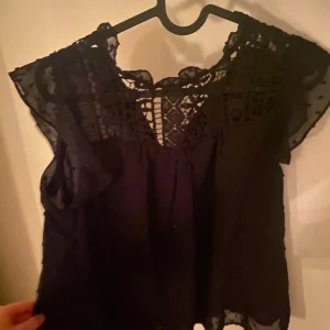Svart blus med spetsdetaljer - Säljer en elegant svart blus med vackra spetsdetaljer vid halsen och ärmarna. Perfekt för både fest och vardag. Blusen har en lös passform och är tillverkad i ett lätt och luftigt material. Ett måste i garderoben för den som älskar stilrena plagg!